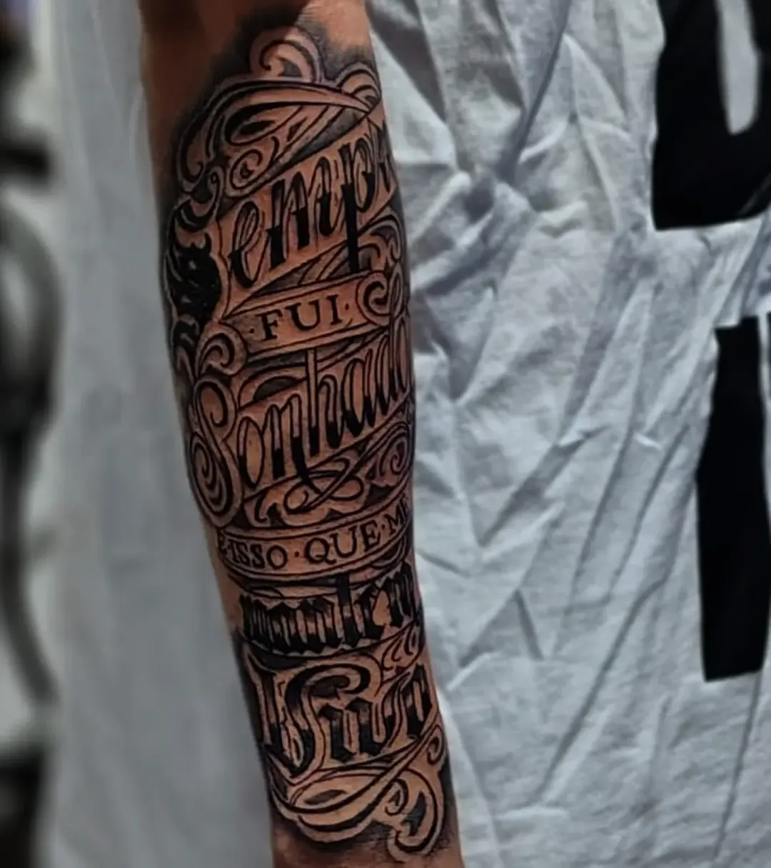 Lettering script con ornamento cubriendo el antebrazo completo, en negro y gris